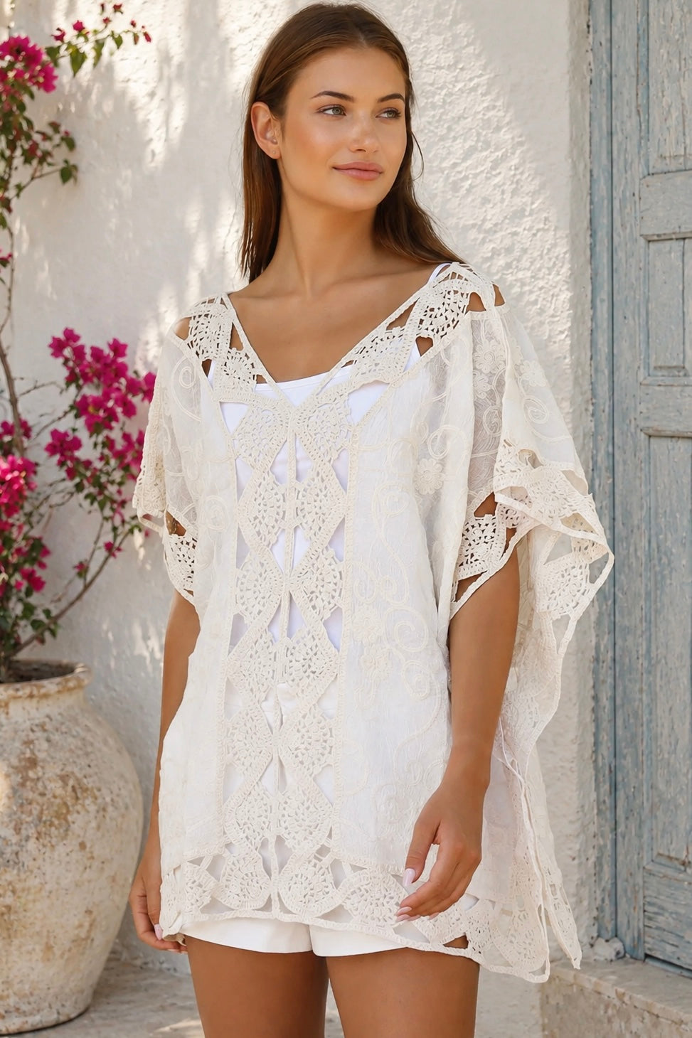 Cream crochet boho kaftan with ornate lace panels and relaxed sleeves in a sunlit Mediterranean coastal setting.