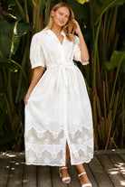 A woman wearing a white lace midi shirt dress with short puff sleeves, a tie waist, and a front slit, standing outdoors in front of tropical green foliage.