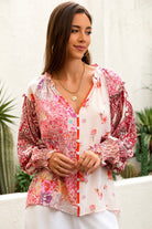 A woman wearing a bohemian-style long-sleeve blouse with a pink, red, and orange floral patchwork print on a white base, styled with white pants and gold jewelry in an outdoor setting.