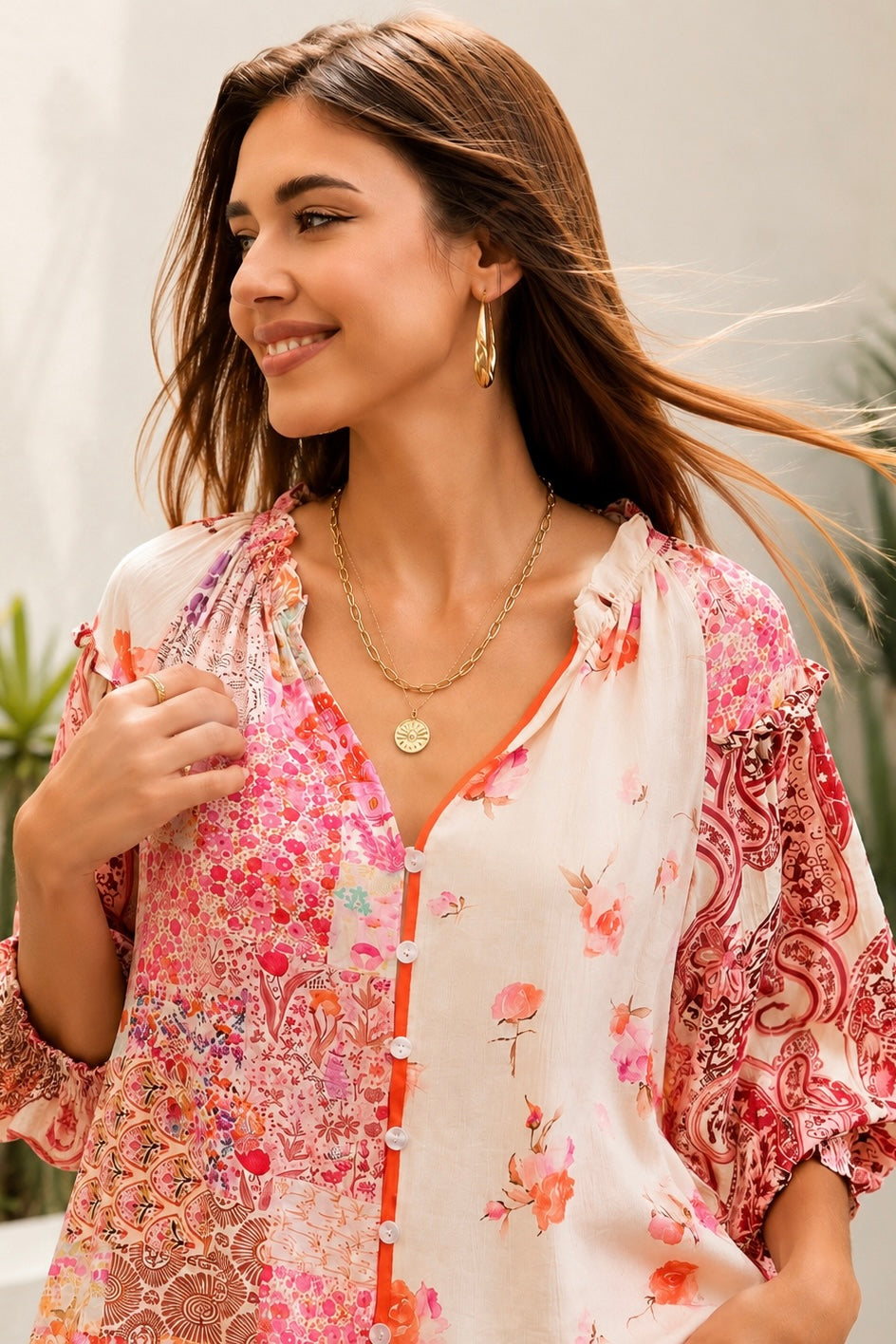 A woman wearing a bohemian-style long-sleeve blouse with a pink, red, and orange floral patchwork print on a white base, styled with white pants and gold jewelry in an outdoor setting.