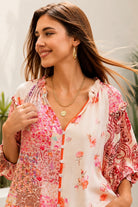 A woman wearing a bohemian-style long-sleeve blouse with a pink, red, and orange floral patchwork print on a white base, styled with white pants and gold jewelry in an outdoor setting.