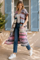 A woman wearing a long, open-front, short-sleeved chunky knit cardigan with horizontal multicolor stripes in pink, blue, beige, brown, and white, paired with a white tank top and blue jeans, standing in front of a window with blue shutters.