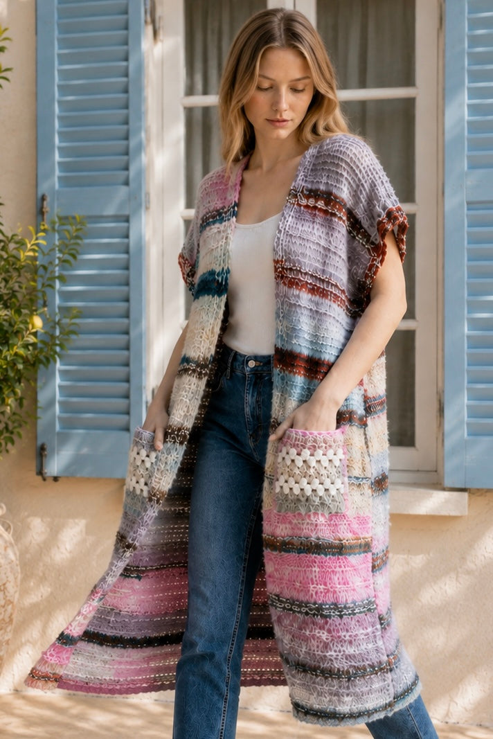 A woman wearing a long, open-front, short-sleeved chunky knit cardigan with horizontal multicolor stripes in pink, blue, beige, brown, and white, paired with a white tank top and blue jeans, standing in front of a window with blue shutters.