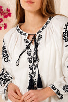 A close-up of a woman wearing a white long-sleeve blouse with black floral embroidery and a black tassel tie at the neckline. Bohemian Love Runway.