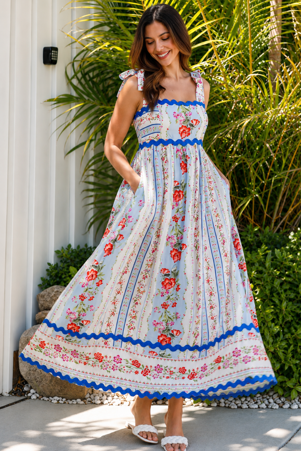 A woman wearing a sleeveless, A-line maxi dress with a white base, blue scalloped trim, and a red and pink floral print, standing outdoors in front of green plants.