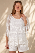 A woman wearing a white lace long-sleeve tunic top with matching white shorts, standing in a sunlit outdoor setting with a beige wall and potted plant in the background.