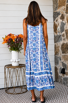 A woman modeling a blue and white patterned maxi dress with spaghetti straps, standing on a patterned tile porch next to a small table with orange flowers.