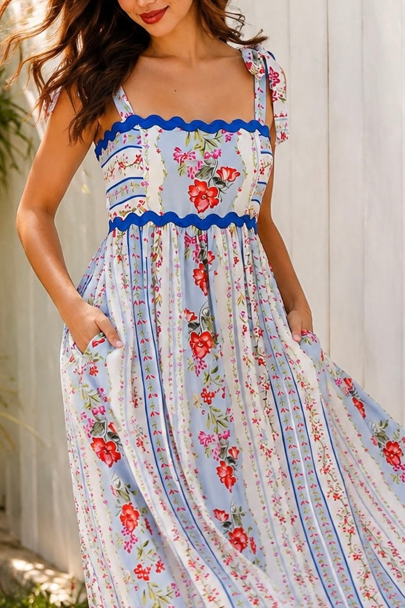 A woman wearing a sleeveless, A-line maxi dress with a white base, blue scalloped trim, and a red and pink floral print, standing outdoors in front of green plants.