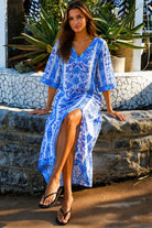 A woman wearing a blue and white patterned kaftan dress with a V-neck and three-quarter sleeves, walking outdoors in front of a stone wall and tropical plants.