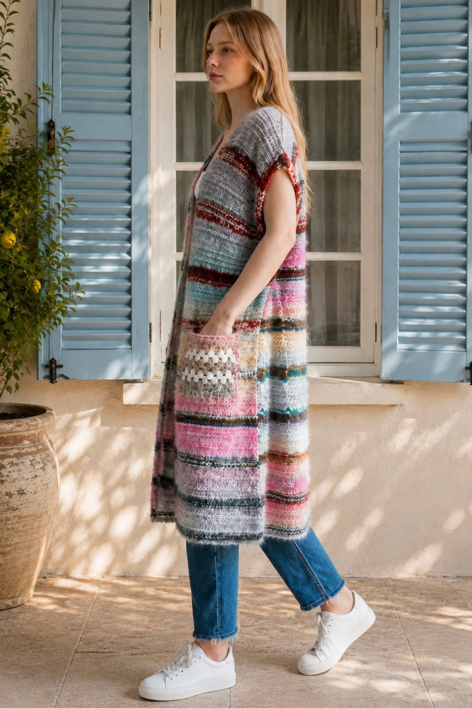 A woman wearing a long, open-front, short-sleeved chunky knit cardigan with horizontal multicolor stripes in pink, blue, beige, brown, and white, paired with a white tank top and blue jeans, standing in front of a window with blue shutters.