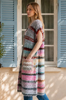 A woman wearing a long, open-front, short-sleeved chunky knit cardigan with horizontal multicolor stripes in pink, blue, beige, brown, and white, paired with a white tank top and blue jeans, standing in front of a window with blue shutters.