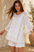 A woman wearing a white lace long-sleeve tunic top with matching white shorts, standing in a sunlit outdoor setting with a beige wall and potted plant in the background.