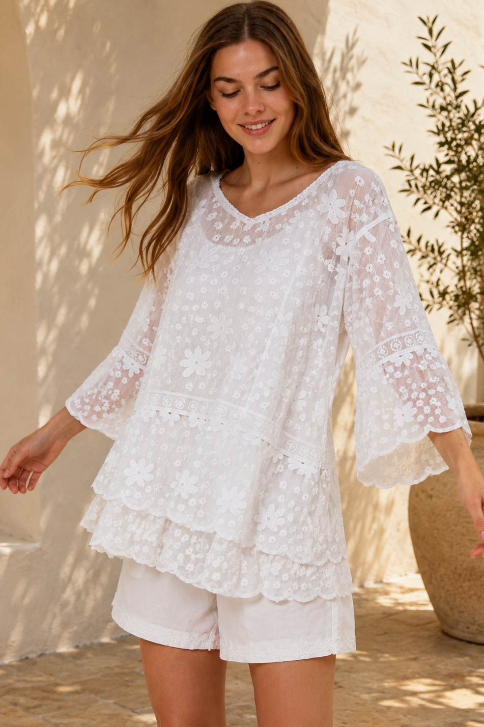 A woman wearing a white lace long-sleeve tunic top with matching white shorts, standing in a sunlit outdoor setting with a beige wall and potted plant in the background.
