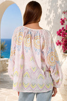 A woman viewed from behind wearing a light pink eyelet blouse with yellow, blue, and green bohemian circular patterns, set against a Mediterranean-style background with a blue sea and white stucco wall.