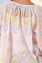 A close-up of a light pink eyelet embroidered blouse with colorful floral and paisley patterns in yellow, blue, green, and purple, featuring a gathered neckline.