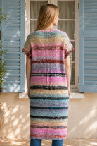 A woman wearing a long, open-front, short-sleeved chunky knit cardigan with horizontal multicolor stripes in pink, blue, beige, brown, and white, paired with a white tank top and blue jeans, standing in front of a window with blue shutters.