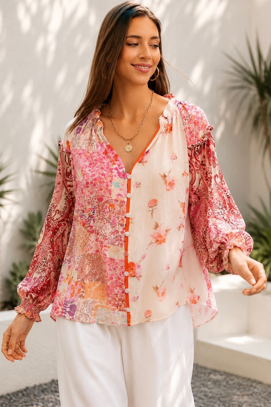 A woman wearing a bohemian-style long-sleeve blouse with a pink, red, and orange floral patchwork print on a white base, styled with white pants and gold jewelry in an outdoor setting.