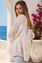 A woman wearing a light pink eyelet lace kimono cardigan with pastel yellow, blue, and green circular patterns, standing outdoors next to a white wall and a potted plant with pink flowers.
