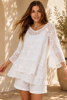 A woman wearing a white lace long-sleeve tunic top with matching white shorts, standing in a sunlit outdoor setting with a beige wall and potted plant in the background.