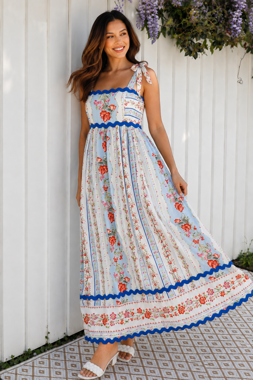 A woman wearing a sleeveless, A-line maxi dress with a white base, blue scalloped trim, and a red and pink floral print, standing outdoors in front of green plants.