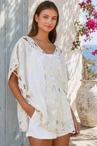 Cream crochet boho kaftan with ornate lace panels and relaxed sleeves in a sunlit Mediterranean coastal setting.