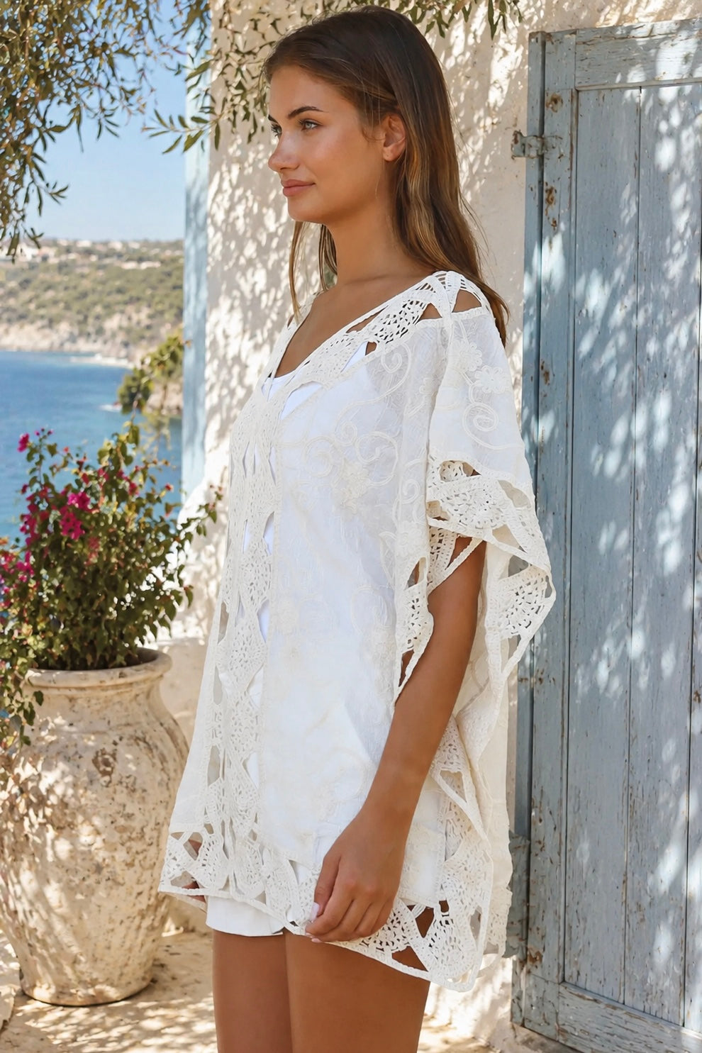 Cream crochet boho kaftan with ornate lace panels and relaxed sleeves in a sunlit Mediterranean coastal setting.