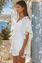 Cream crochet boho kaftan with ornate lace panels and relaxed sleeves in a sunlit Mediterranean coastal setting.
