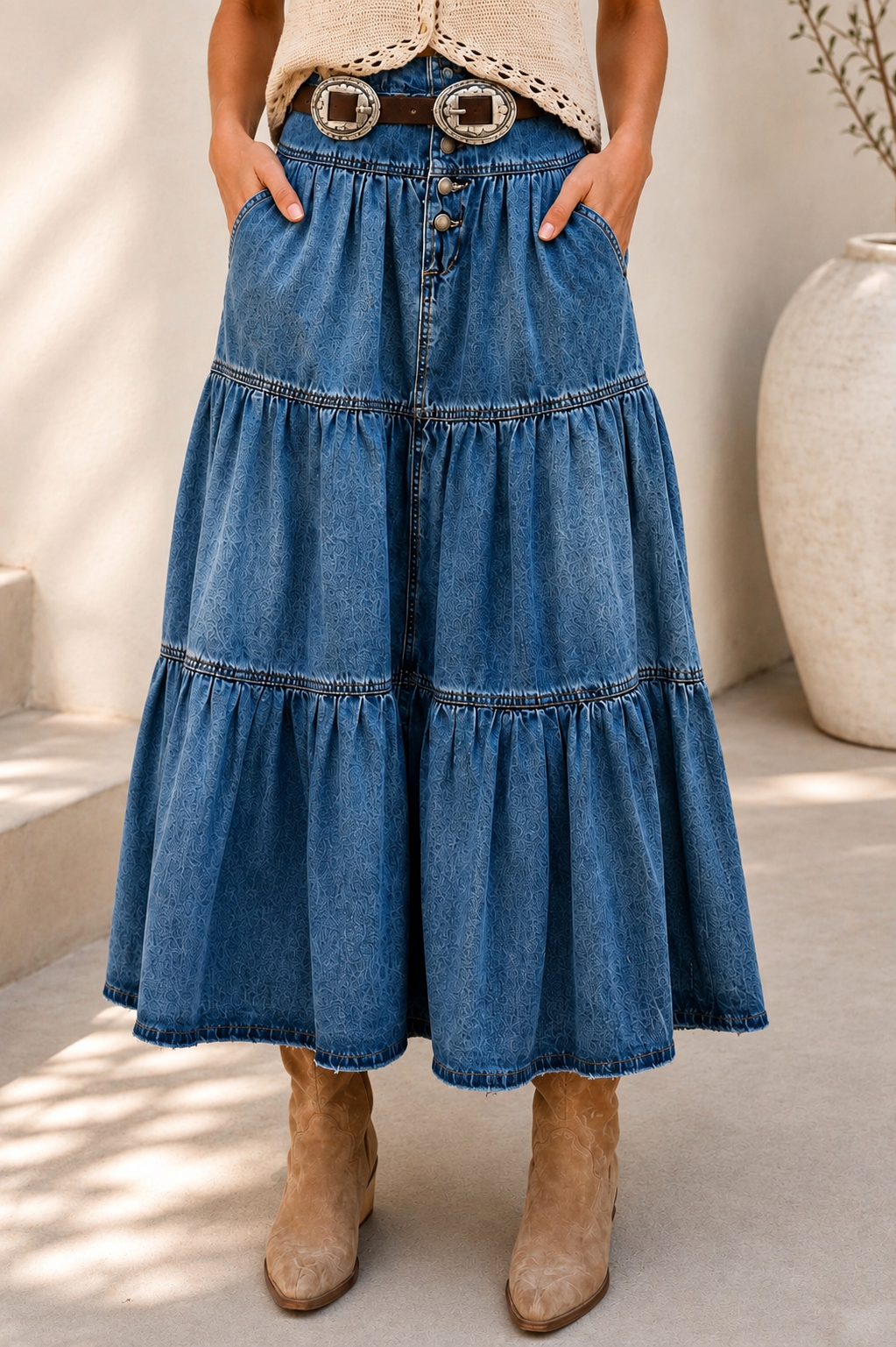 A woman models a bohemian-style outfit featuring a high-waisted, tiered blue denim skirt with a decorative belt, paired with a cream crochet top and beige boots, set against a sunlit neutral background.