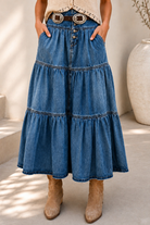 A woman models a bohemian-style outfit featuring a high-waisted, tiered blue denim skirt with a decorative belt, paired with a cream crochet top and beige boots, set against a sunlit neutral background.