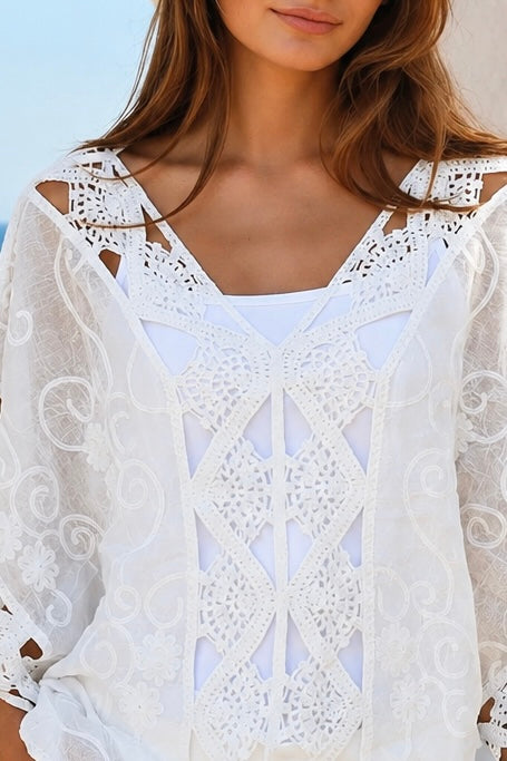 A woman wearing a white eyelet lace cover-up top with cutout details, standing outdoors in front of a white stucco wall with a blue ocean and pink flowers in the background.