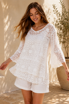 A woman wearing a white lace long-sleeve tunic top with matching white shorts, standing in a sunlit outdoor setting with a beige wall and potted plant in the background.