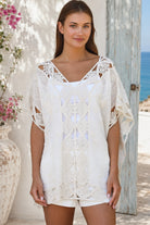 Cream crochet boho kaftan with ornate lace panels and relaxed sleeves in a sunlit Mediterranean coastal setting.