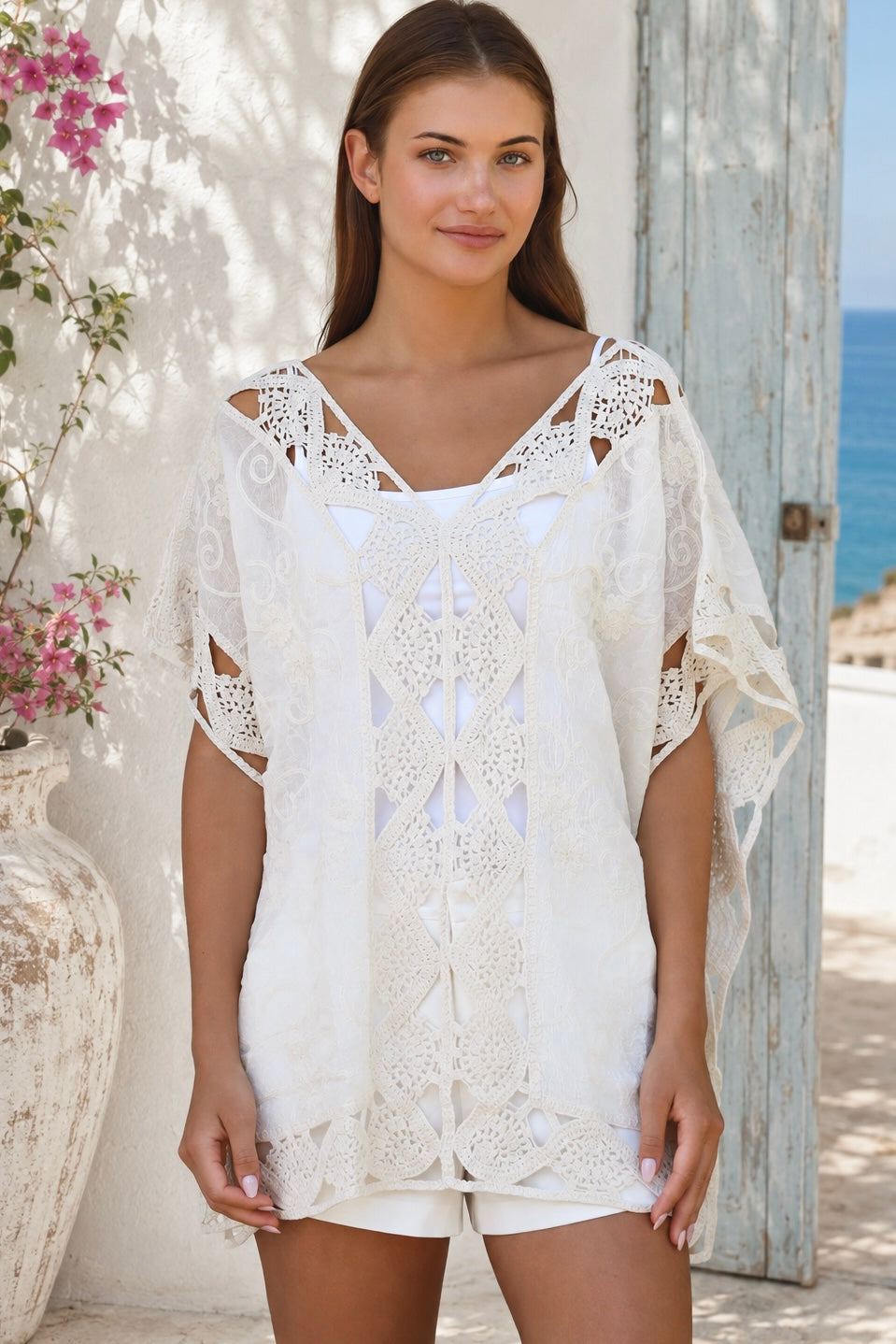 Cream crochet boho kaftan with ornate lace panels and relaxed sleeves in a sunlit Mediterranean coastal setting.