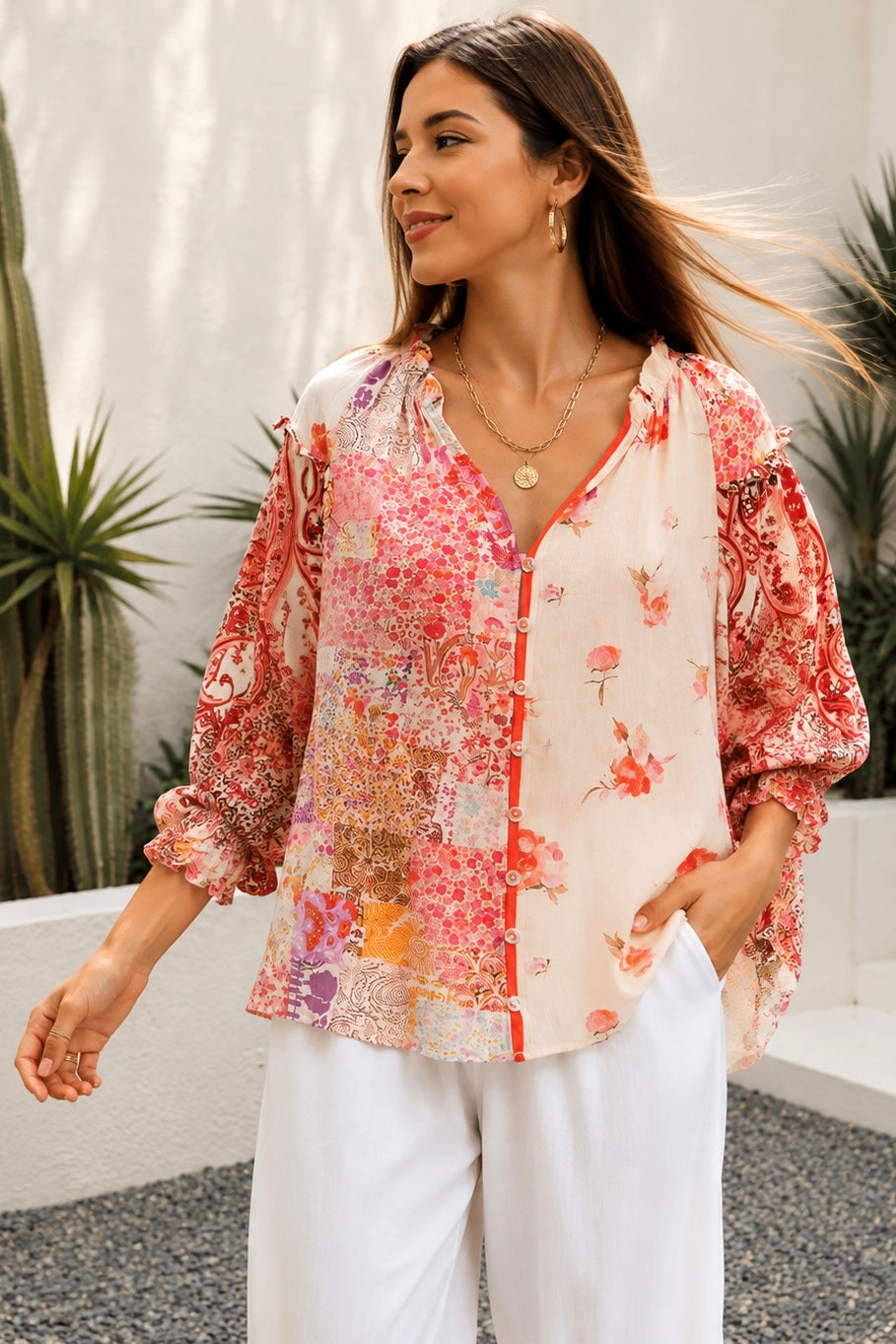 A woman wearing a bohemian-style long-sleeve blouse with a pink, red, and orange floral patchwork print on a white base, styled with white pants and gold jewelry in an outdoor setting.