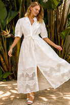 A woman wearing a white lace midi shirt dress with short puff sleeves, a tie waist, and a front slit, standing outdoors in front of tropical green foliage.