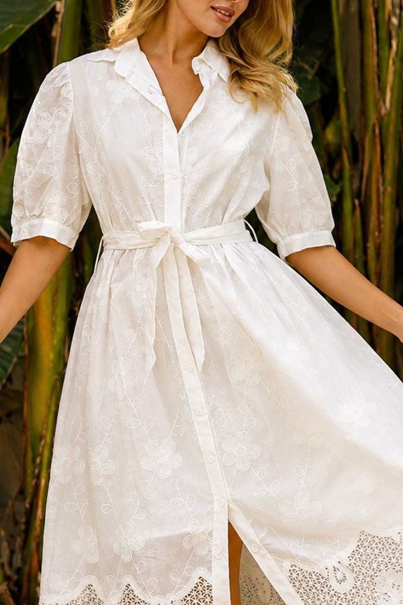 A woman wearing a white lace midi shirt dress with short puff sleeves, a tie waist, and a front slit, standing outdoors in front of tropical green foliage.