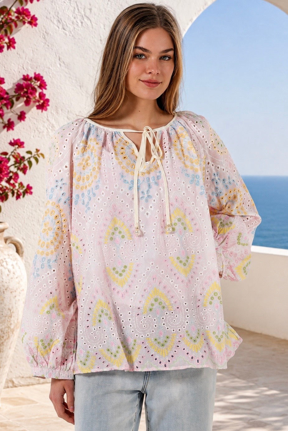A woman wearing a light pink eyelet blouse with a multicolor pastel geometric pattern, featuring a tie-neck detail and long sleeves, paired with light-wash jeans in an outdoor setting with a sea view background.