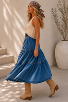 A woman models a bohemian-style outfit featuring a high-waisted, tiered blue denim skirt with a decorative belt, paired with a cream crochet top and beige boots, set against a sunlit neutral background.