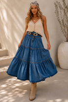 A woman models a bohemian-style outfit featuring a high-waisted, tiered blue denim skirt with a decorative belt, paired with a cream crochet top and beige boots, set against a sunlit neutral background.