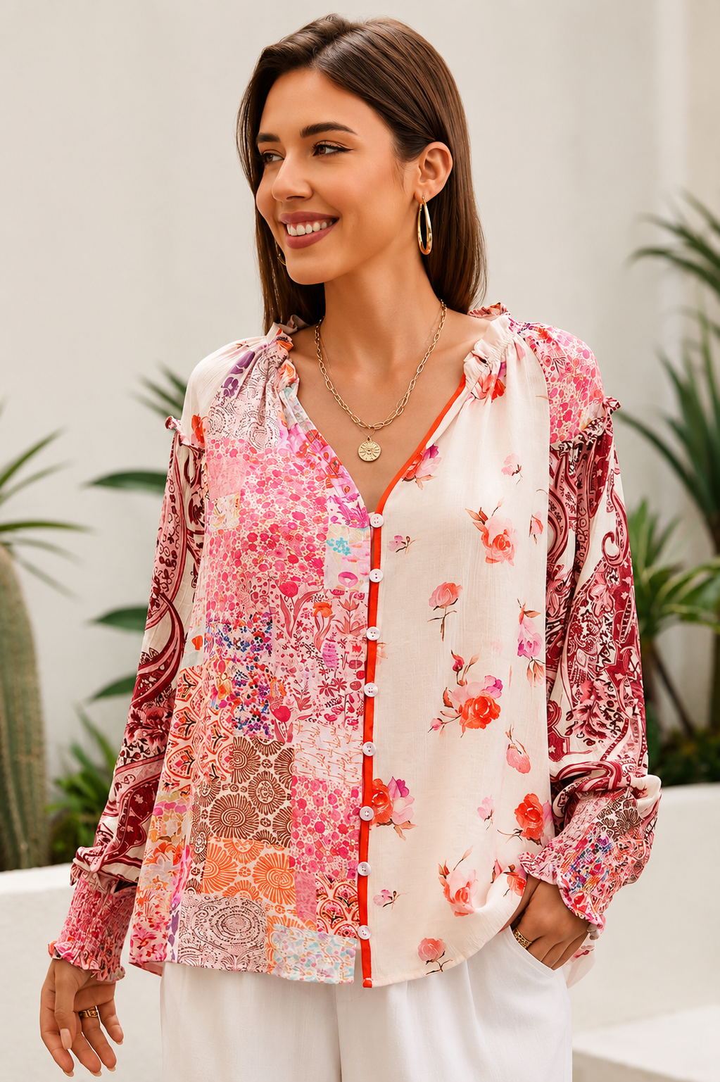 A woman wearing a bohemian-style long-sleeve blouse with a pink, red, and orange floral patchwork print on a white base, styled with white pants and gold jewelry in an outdoor setting.