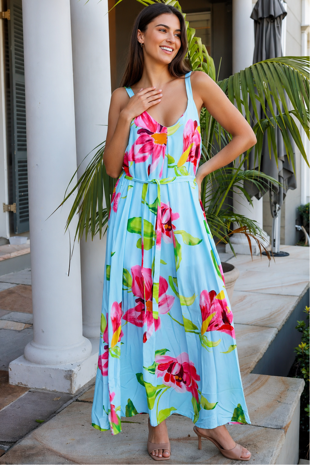 A woman wearing a light blue sleeveless maxi dress with a vibrant pink and green floral print, standing on a stone patio in front of white columns and tropical plants.