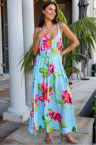 A woman wearing a light blue sleeveless maxi dress with a vibrant pink and green floral print, standing on a stone patio in front of white columns and tropical plants.