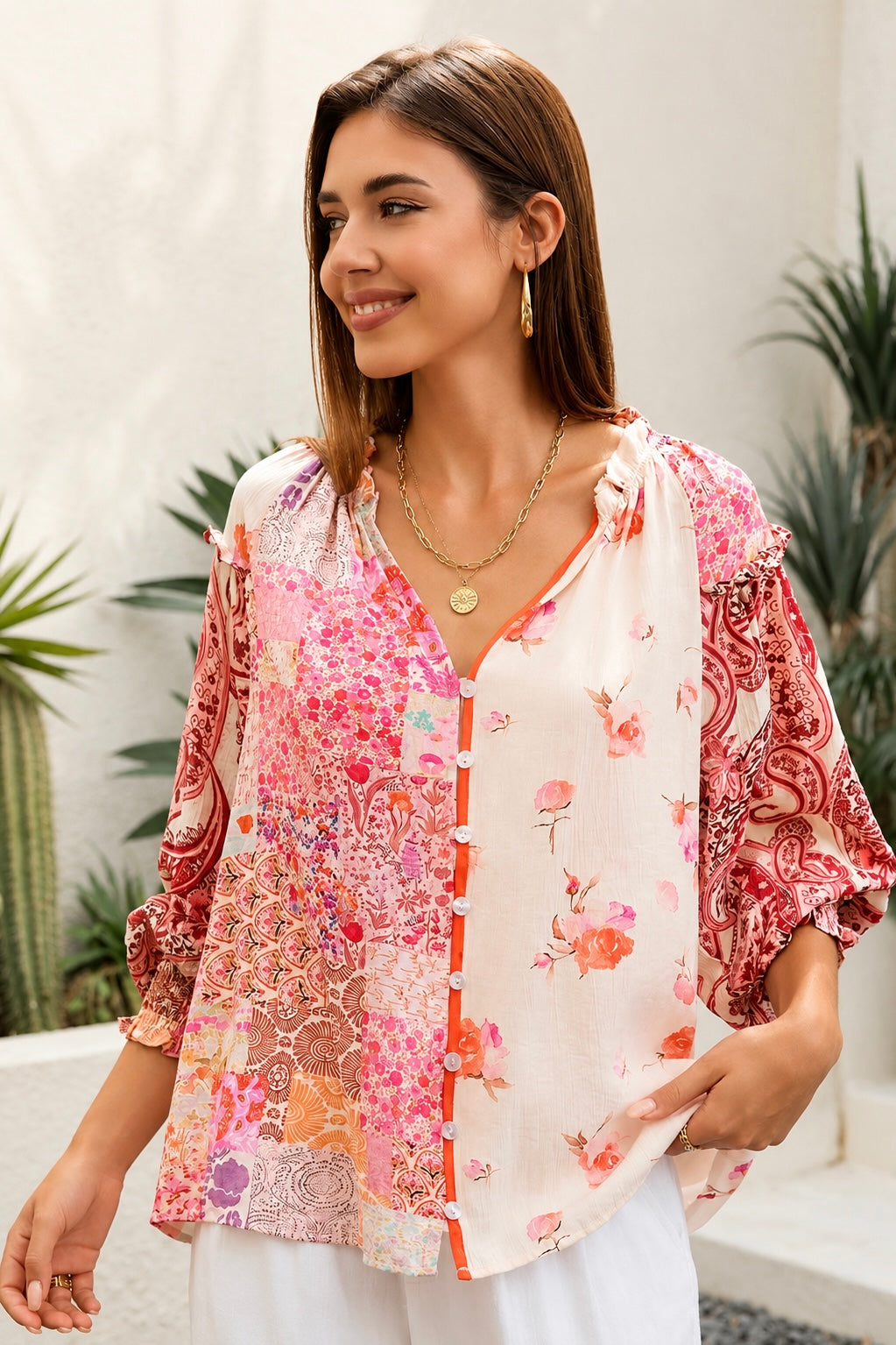 A woman wearing a bohemian-style long-sleeve blouse with a pink, red, and orange floral patchwork print on a white base, styled with white pants and gold jewelry in an outdoor setting.