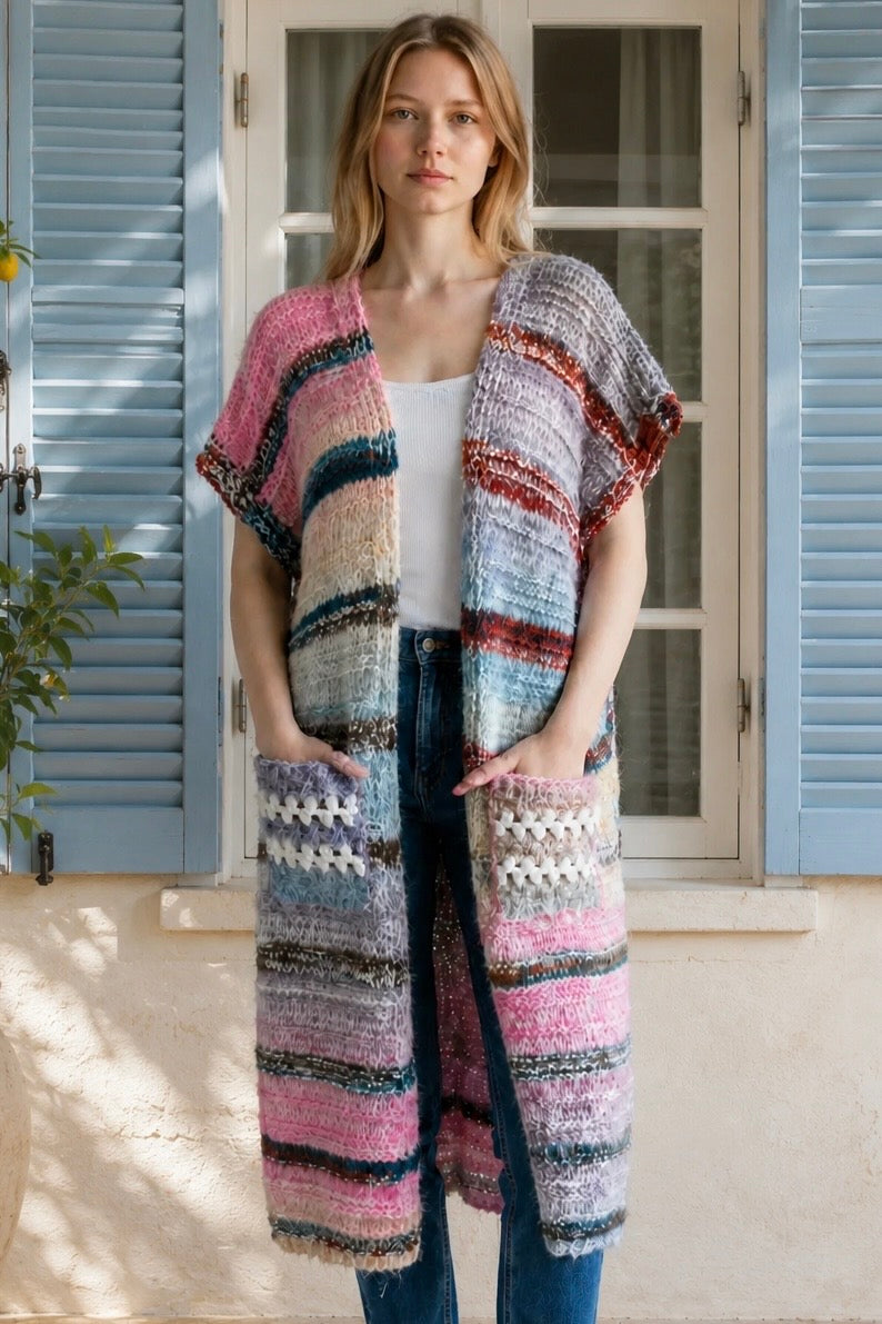 A woman wearing a long, open-front, short-sleeved chunky knit cardigan with horizontal multicolor stripes in pink, blue, beige, brown, and white, paired with a white tank top and blue jeans, standing in front of a window with blue shutters.