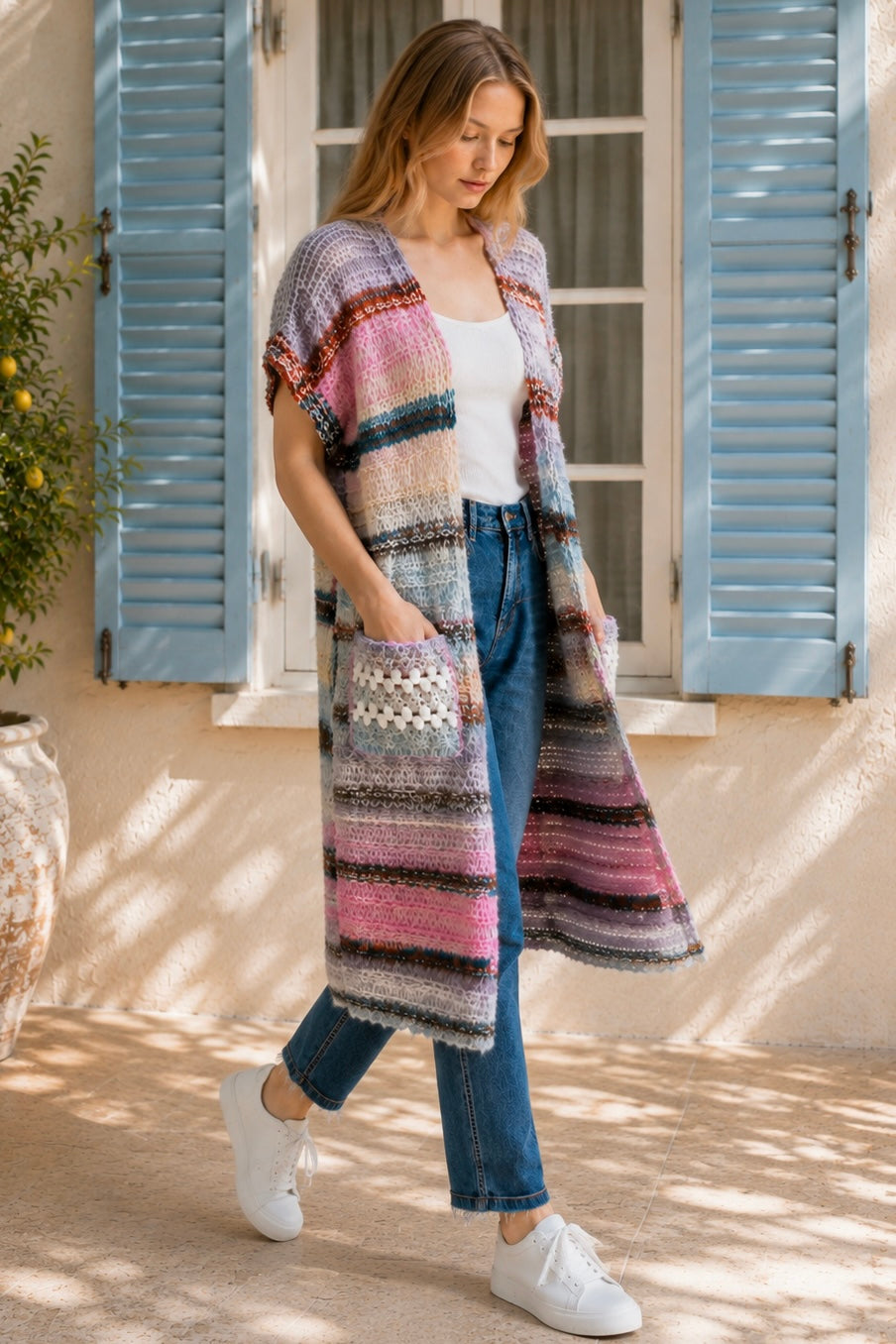 A woman wearing a long, open-front, short-sleeved chunky knit cardigan with horizontal multicolor stripes in pink, blue, beige, brown, and white, paired with a white tank top and blue jeans, standing in front of a window with blue shutters.