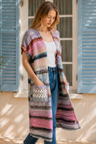 A woman wearing a long, open-front, short-sleeved chunky knit cardigan with horizontal multicolor stripes in pink, blue, beige, brown, and white, paired with a white tank top and blue jeans, standing in front of a window with blue shutters.