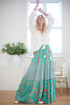Woman wearing a white blouse and a long turquoise floral skirt in a bright room.
