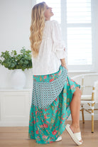 Woman wearing a white blouse and a long turquoise floral skirt in a bright room.
