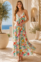 A woman modeling a sleeveless, tiered maxi dress with a vibrant multicolor floral print on a cream background, set in a sunlit Mediterranean-style patio with a sea view in the background.