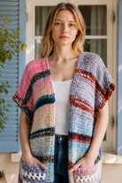 A woman wearing a long, open-front, short-sleeved chunky knit cardigan with horizontal multicolor stripes in pink, blue, beige, brown, and white, paired with a white tank top and blue jeans, standing in front of a window with blue shutters.